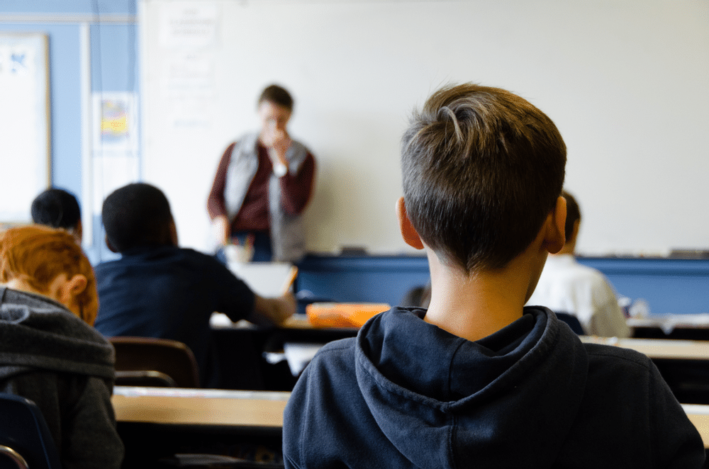 The back of a students head as faces the classroom whiteboard and his male teacher lecturing in front of it,