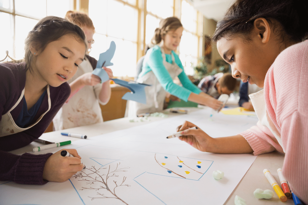 Two young female students lean over a large piece of paper in order to collaborate drawing a picture.