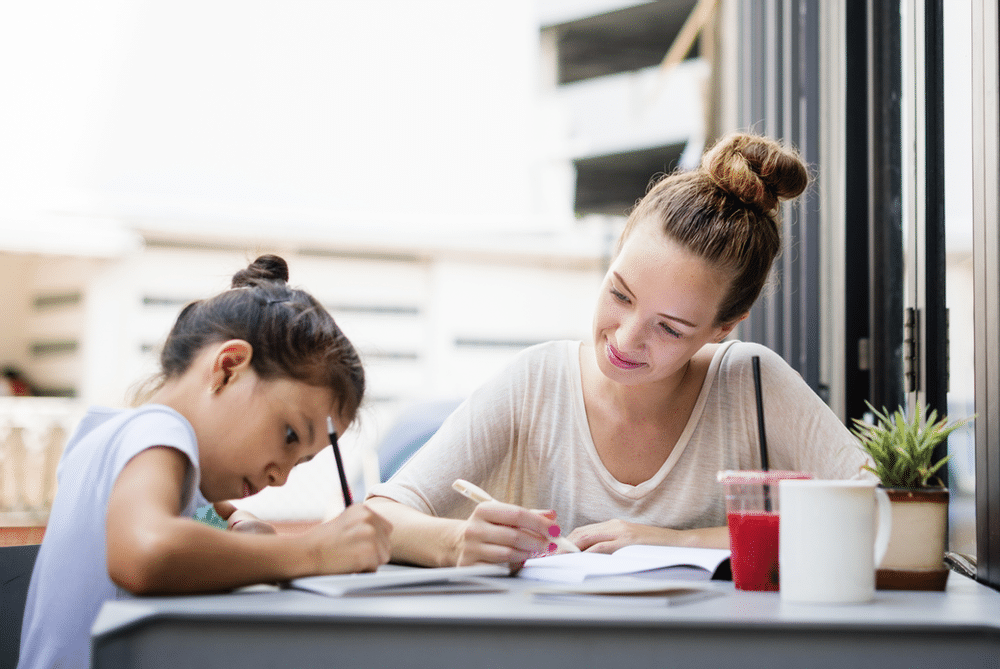 A female-presenting elementary-age child writes in a book while leaning at a table outdoors. A female-presenting adult watches her writing with a smile.