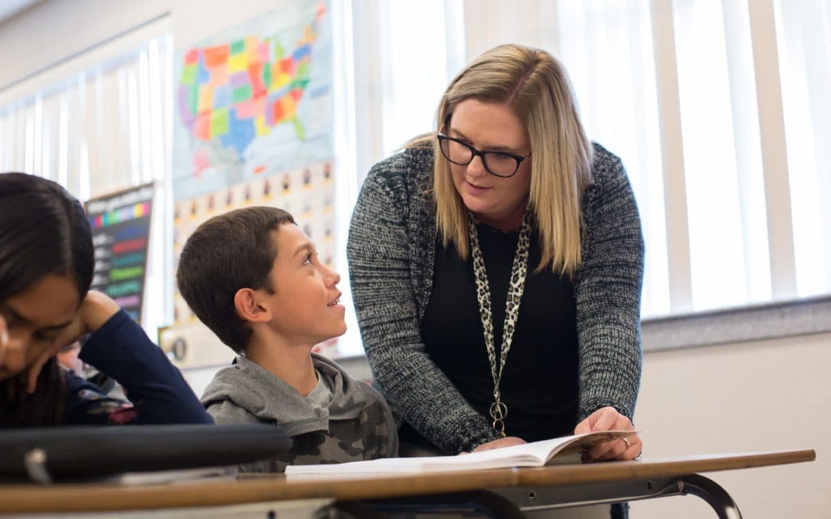 2 students seated in a classroom, conversing with their teacher over a book