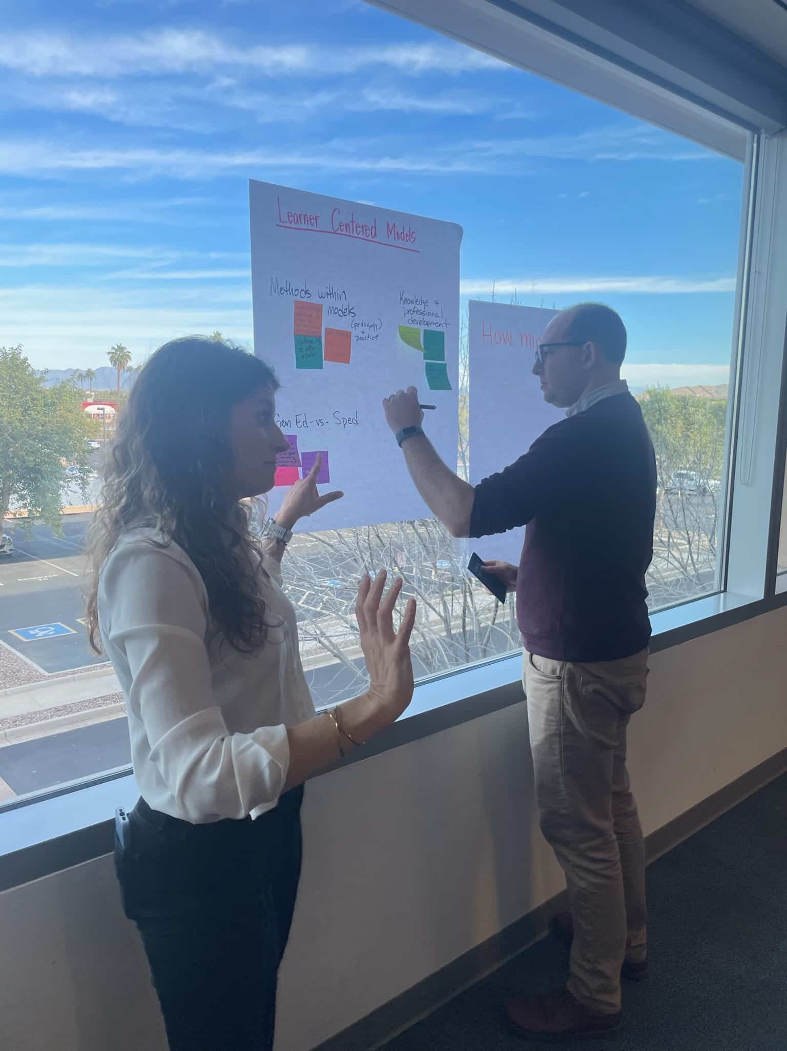 Two people stand by a large window, discussing ideas in front of a whiteboard filled with notes and diagrams.