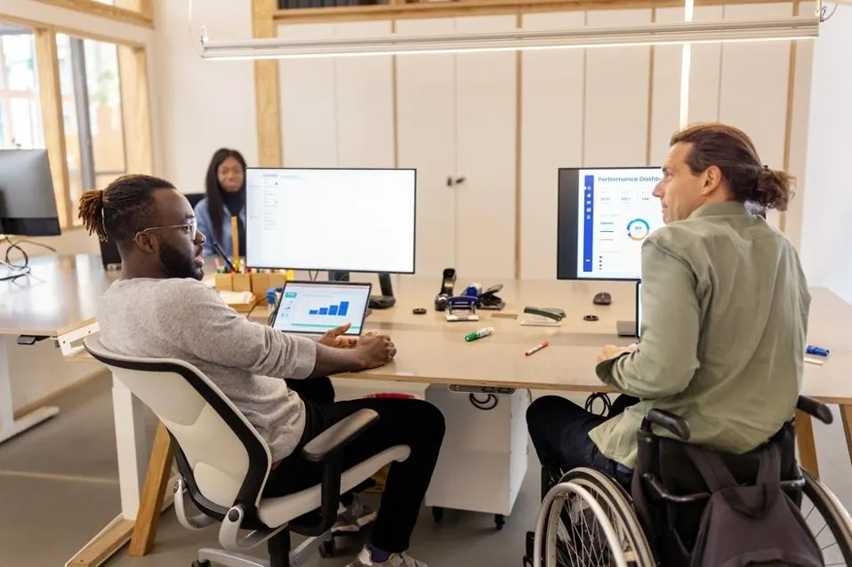 A man sitting in a chair and a man sitting in a wheel chair at their desk looking at computer monitors
