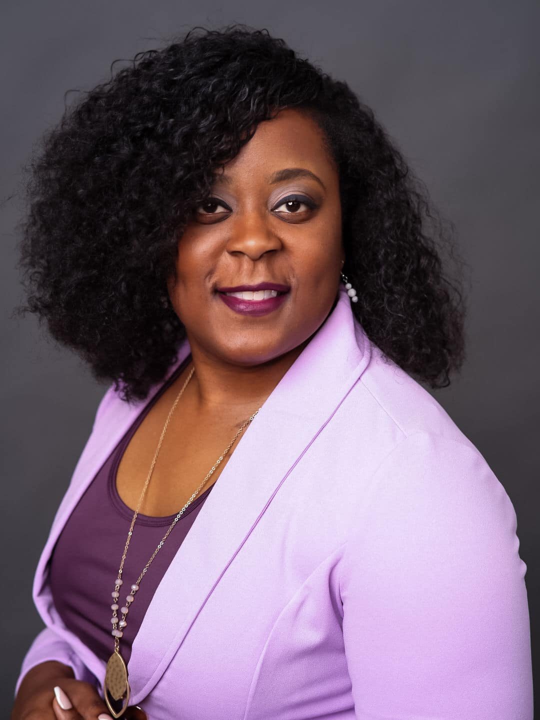 A woman with curly hair wearing a purple blazer, smiling confidently at the camera.
